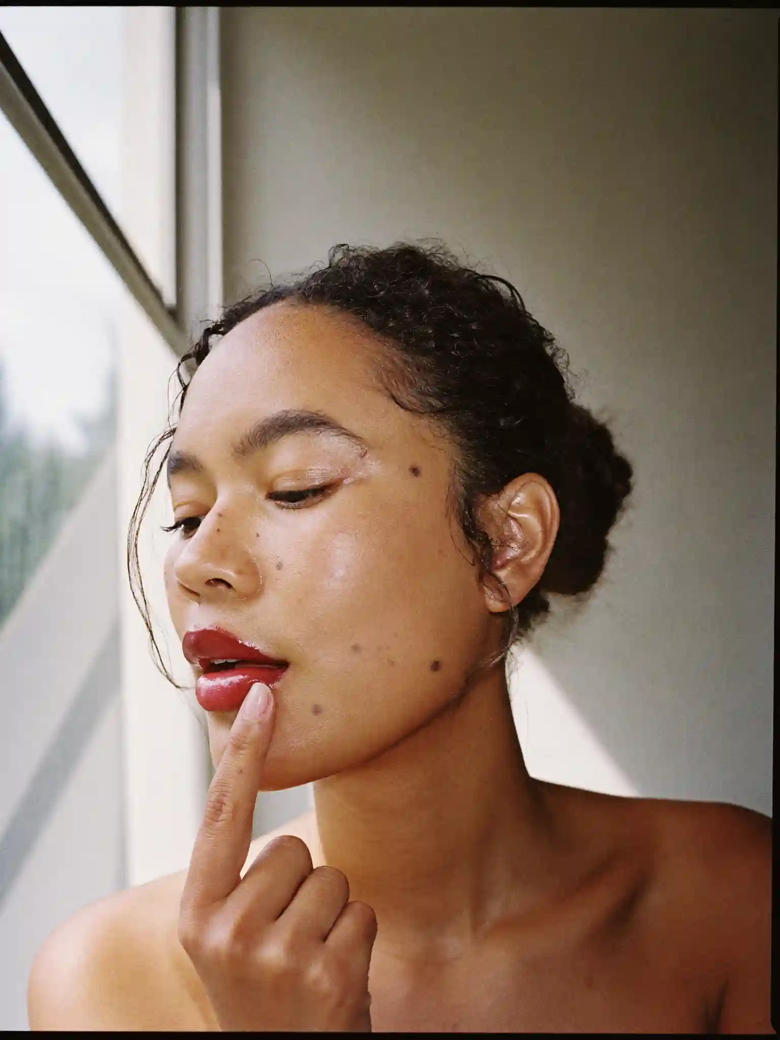 A woman pressing deep red lip colour into her lower lip with a fingertip, lit by window light.