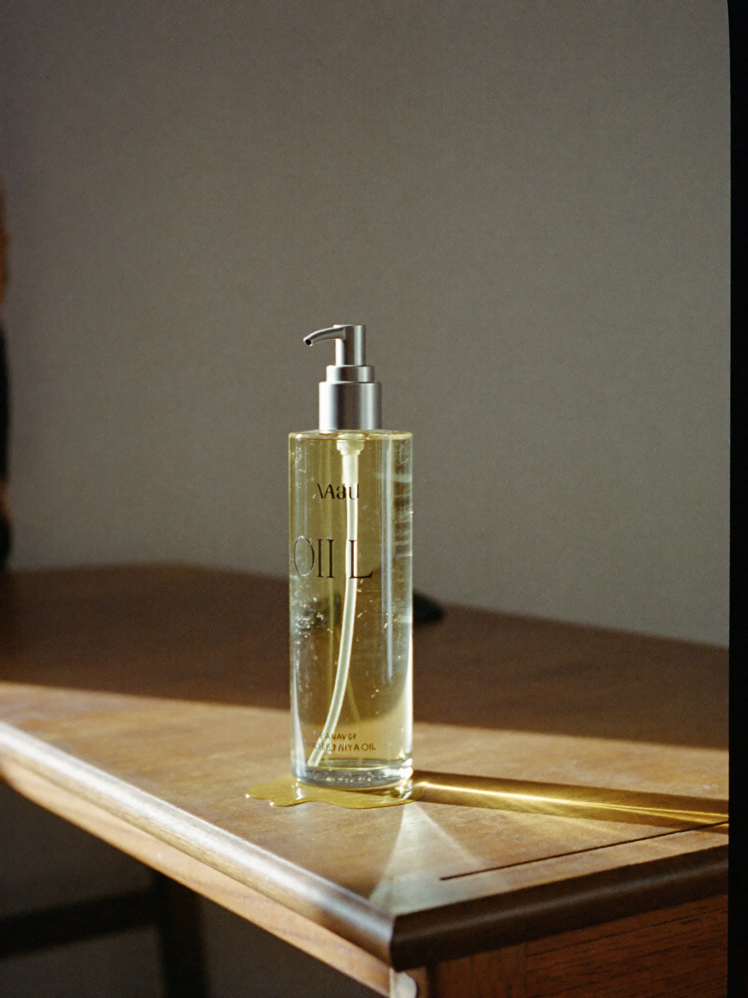 A tall glass bottle of body oil with a metal pump, beside a small pool of oil.