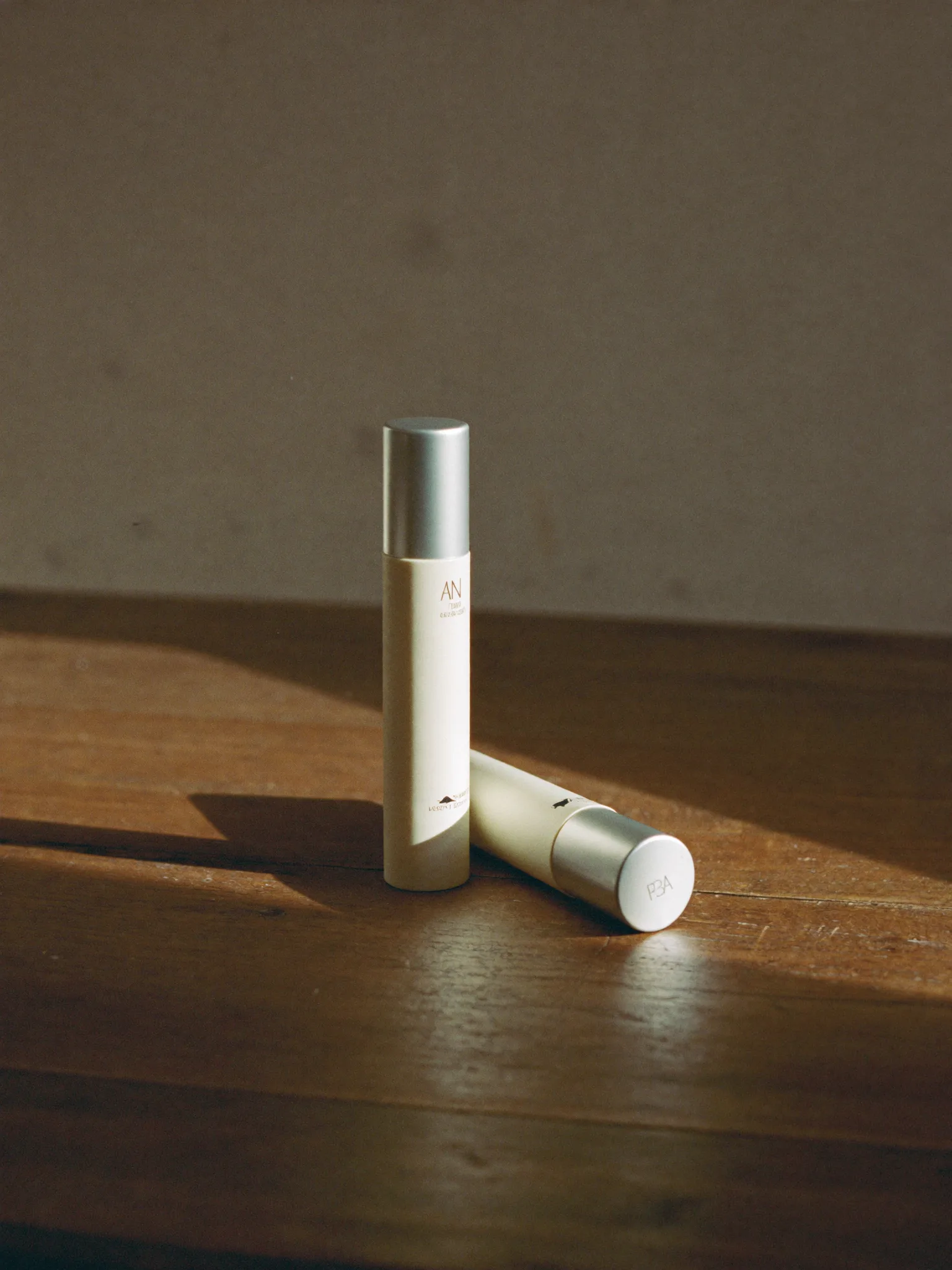 Two cream-coloured lip lacquer tubes on a wooden table in afternoon light.