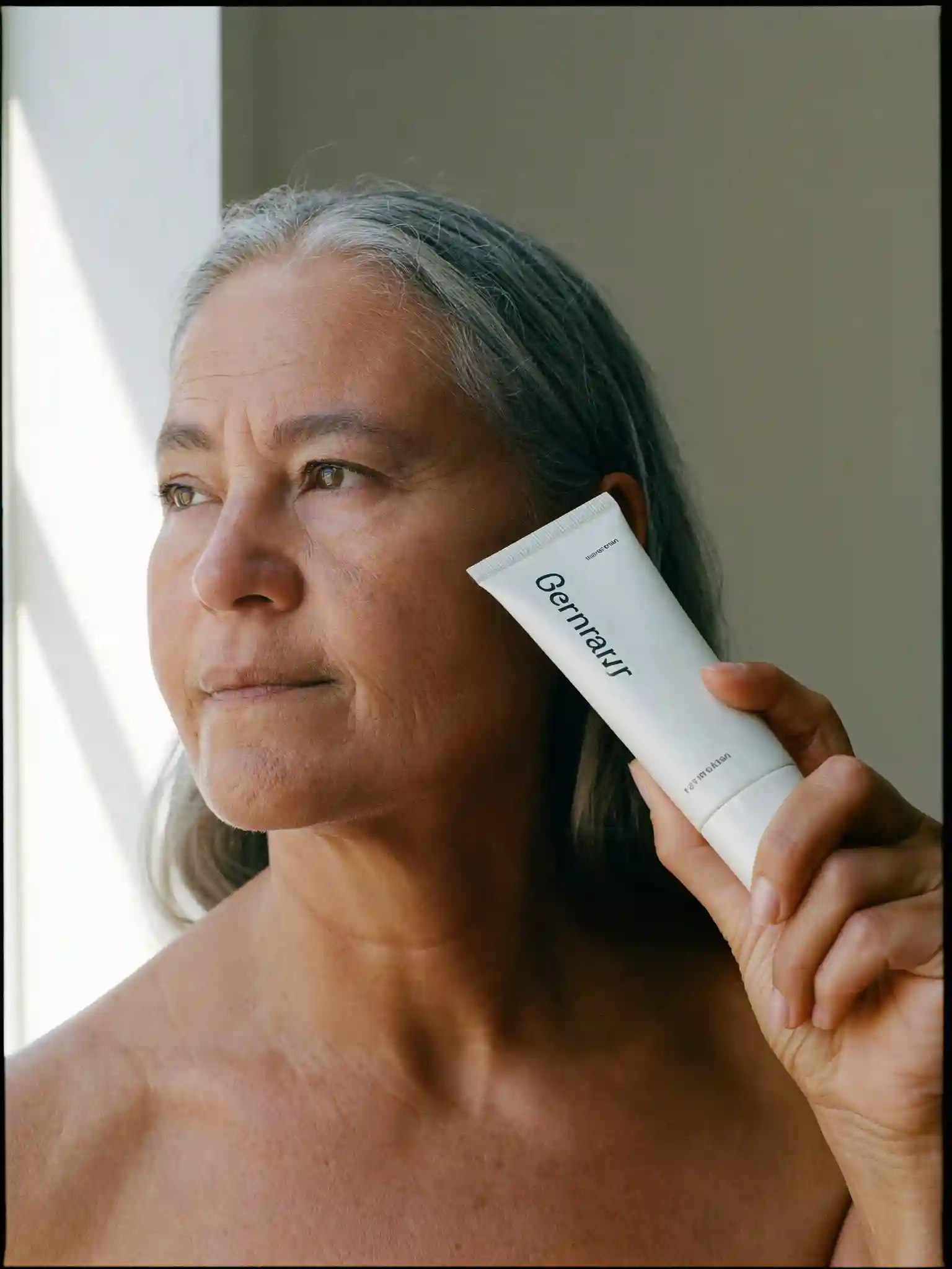 A woman lifting a small unlabelled tube toward her cheek, lit by window light.
